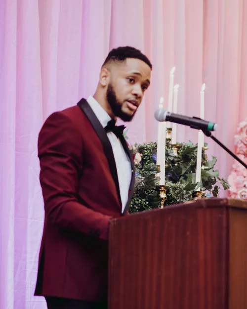 Javon Webster, in a burgundy tuxedo, speaking at a podium.